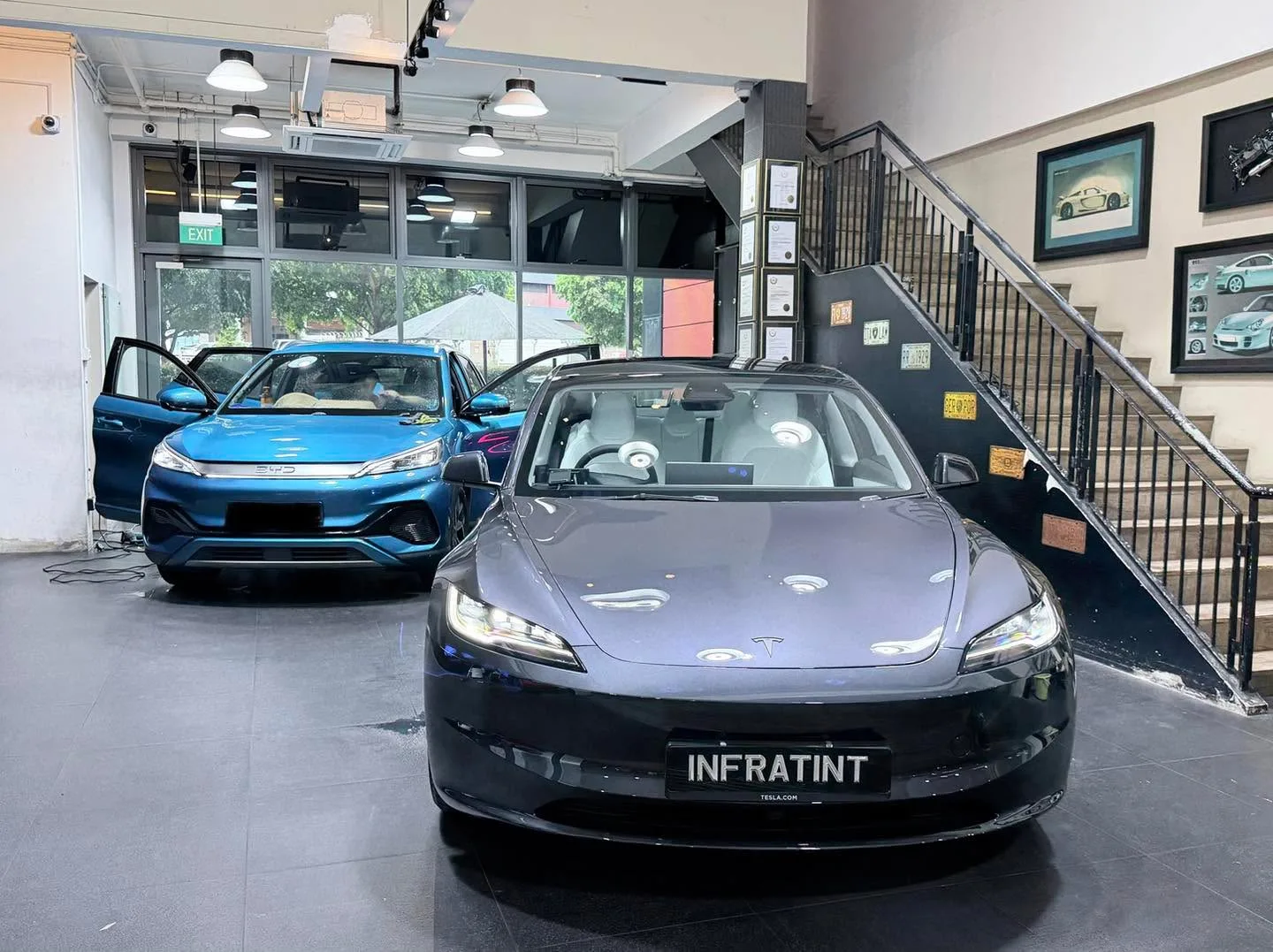 Blue BYD Atto 3 with open door next to grey Tesla sedan indoors.