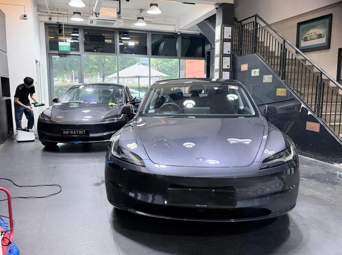 A dark grey Tesla Model 3 and dark grey Tesla sedan inside the workshop near large windows.