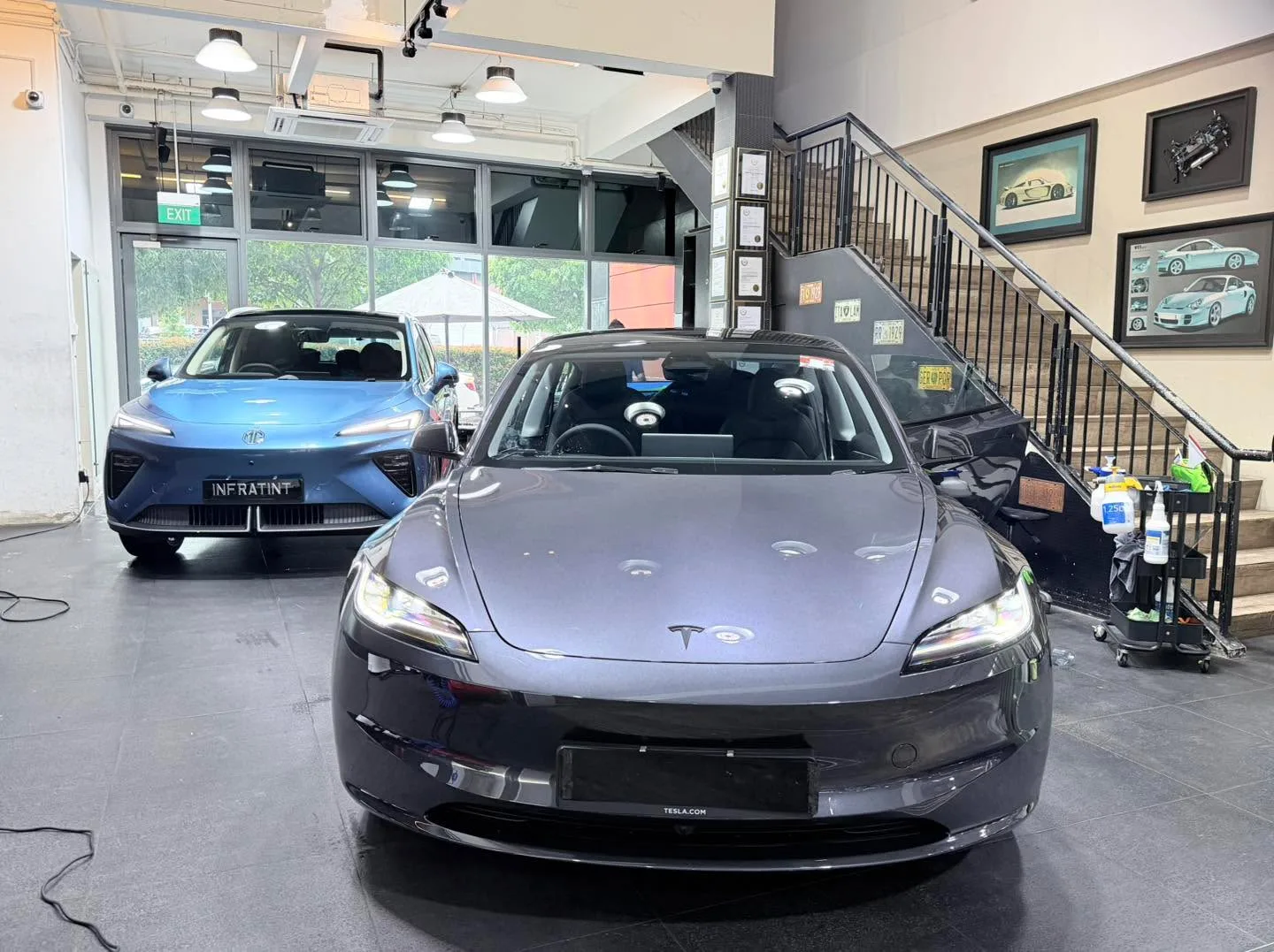 A black Lexus NX with mesh grille in the foreground and white Tesla sedan behind.
