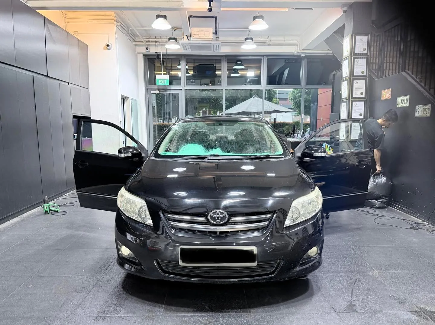A black Toyota Altis saloon with doors open, viewed from the side inside the workshop.