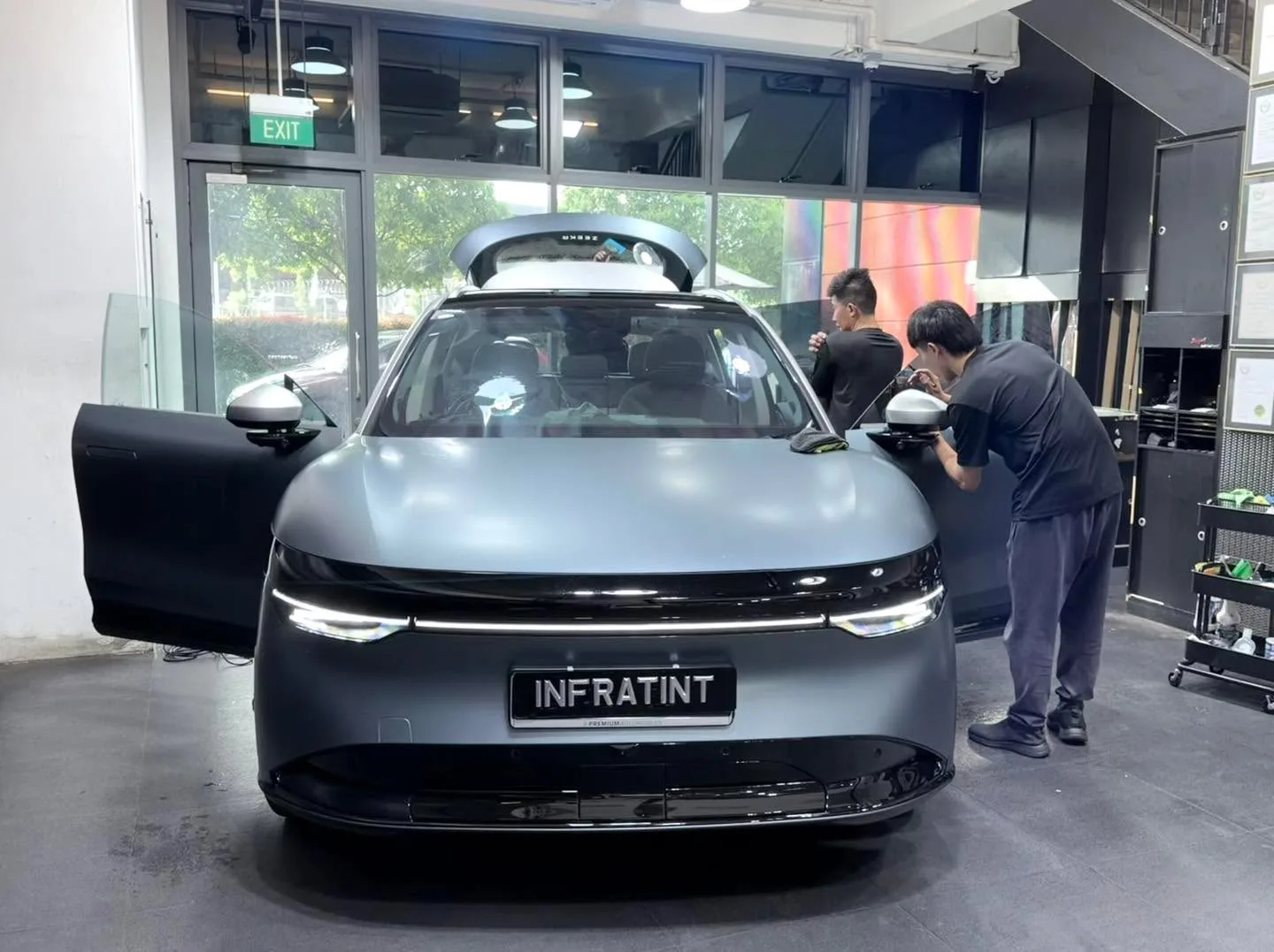 A grey electric saloon with doors open, technicians working on windows near the entrance.
