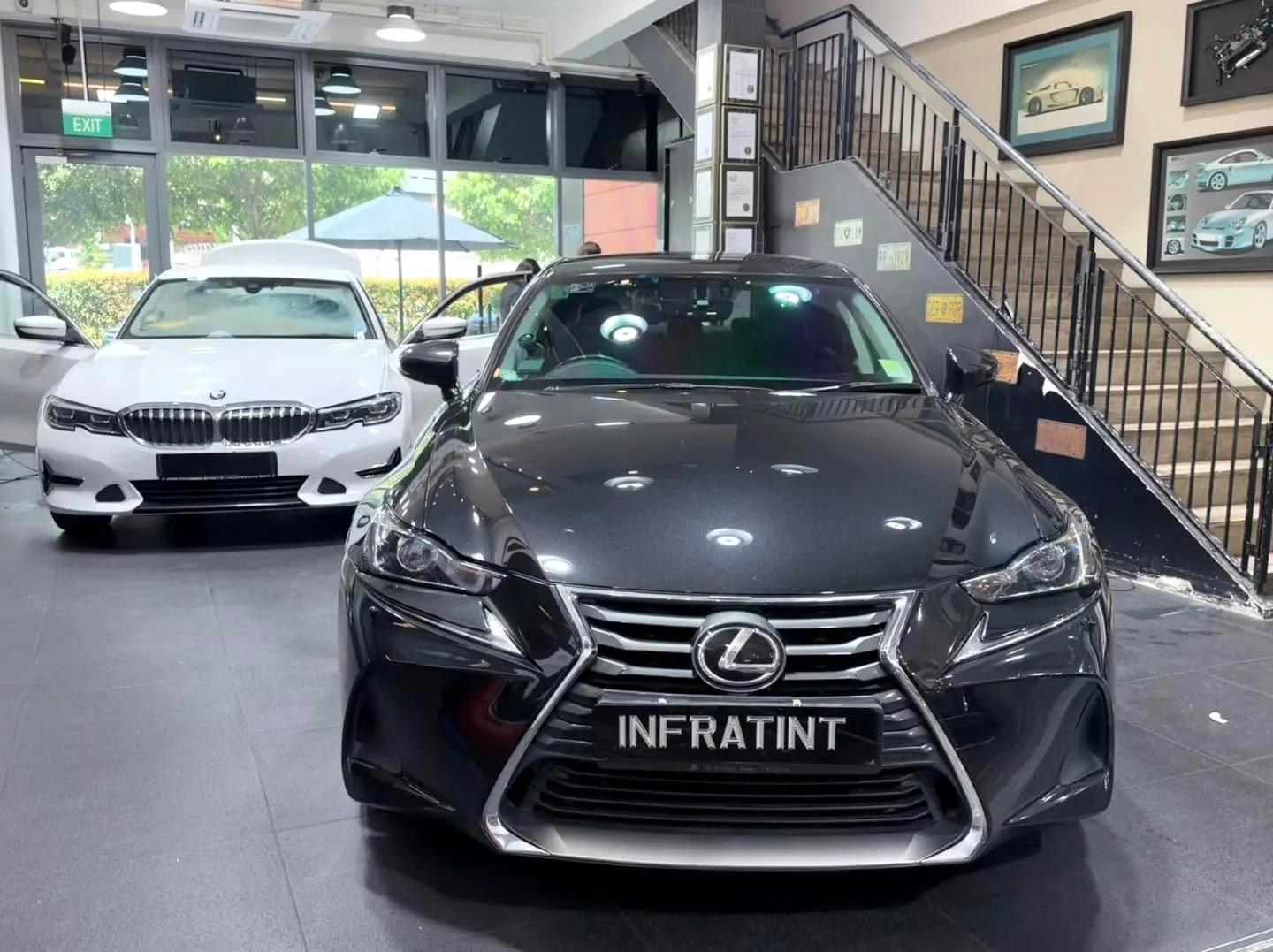 A black Lexus IS sedan with Infratint branding, viewed from the front, with a white BMW visible behind it.