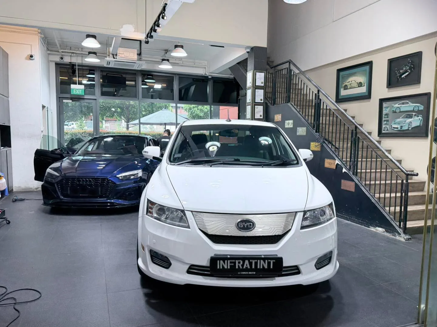 A white BYD sedan viewed from the front at the workshop, with a dark blue car visible in the background.