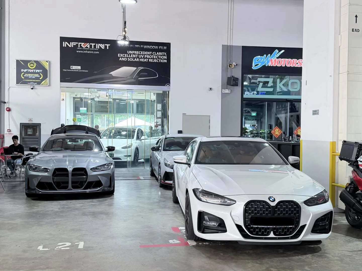A white BMW M4 in the foreground and a grey BMW sedan in the background, parked in an Infratint workshop.