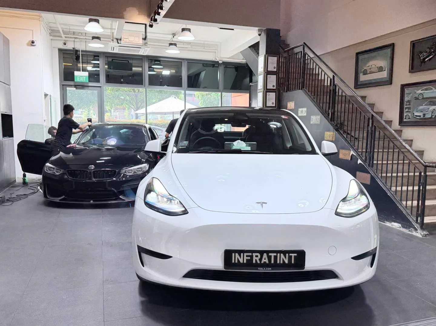 A white Tesla sedan facing the camera, with a black BMW coupe to the left, both in workshop.