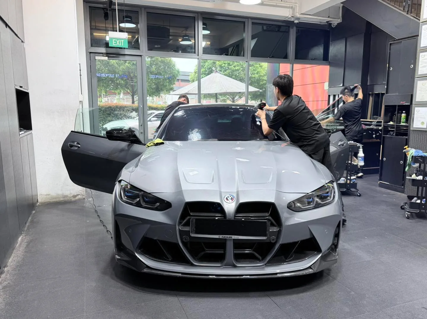 A white BMW M4 facing forwards with its door open, technicians applying film to the windows.