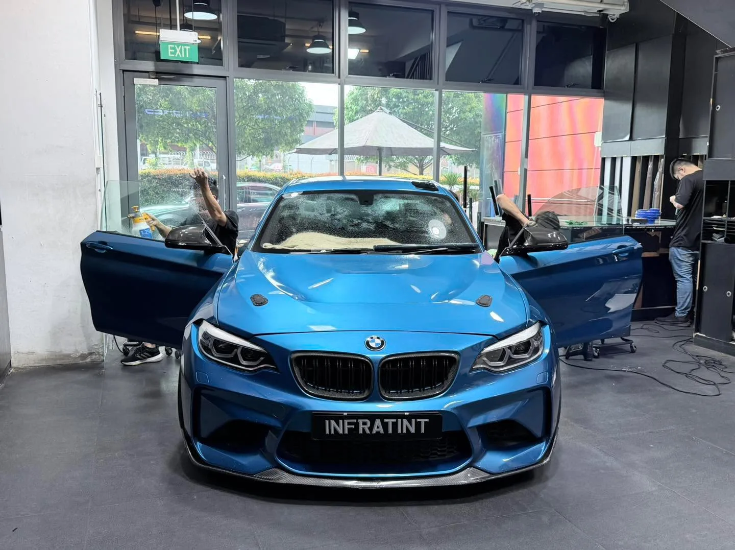 A bright blue BMW M2 with both doors open, viewed head-on in the workshop.