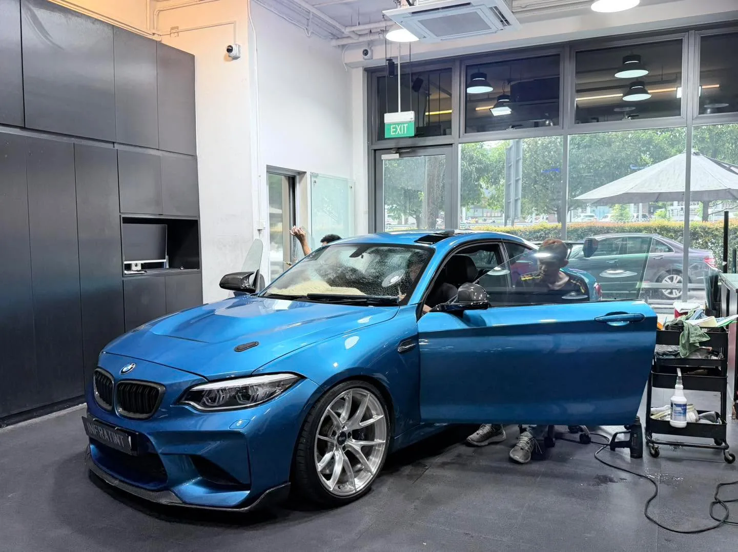 A bright blue BMW M2 coupe with its driver door open during window film application.