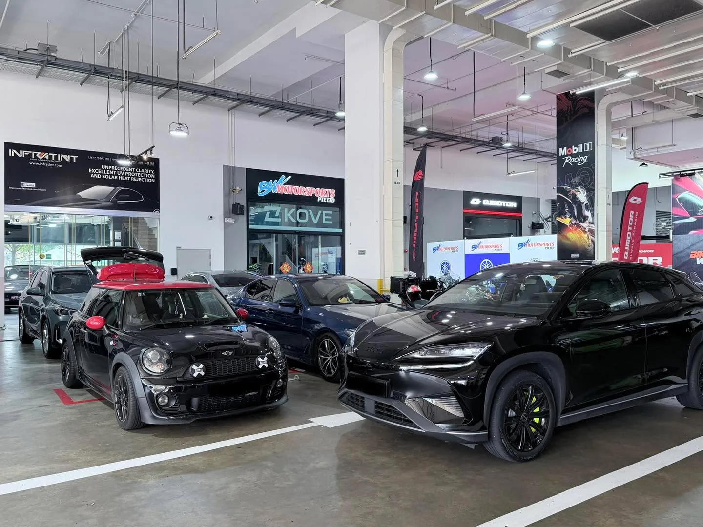 A black Mini JCW with red accents and a blue BMW parked indoors at a workshop.