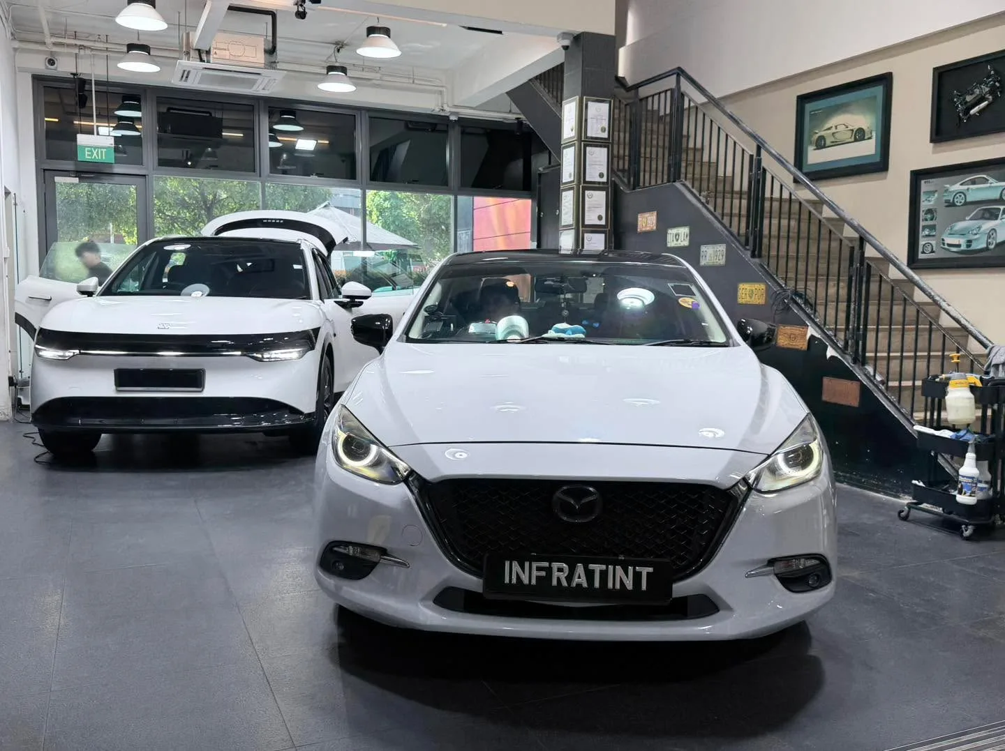 A white Mazda 3 hatchback in the foreground with its windows tinted, viewed from the front at the workshop.