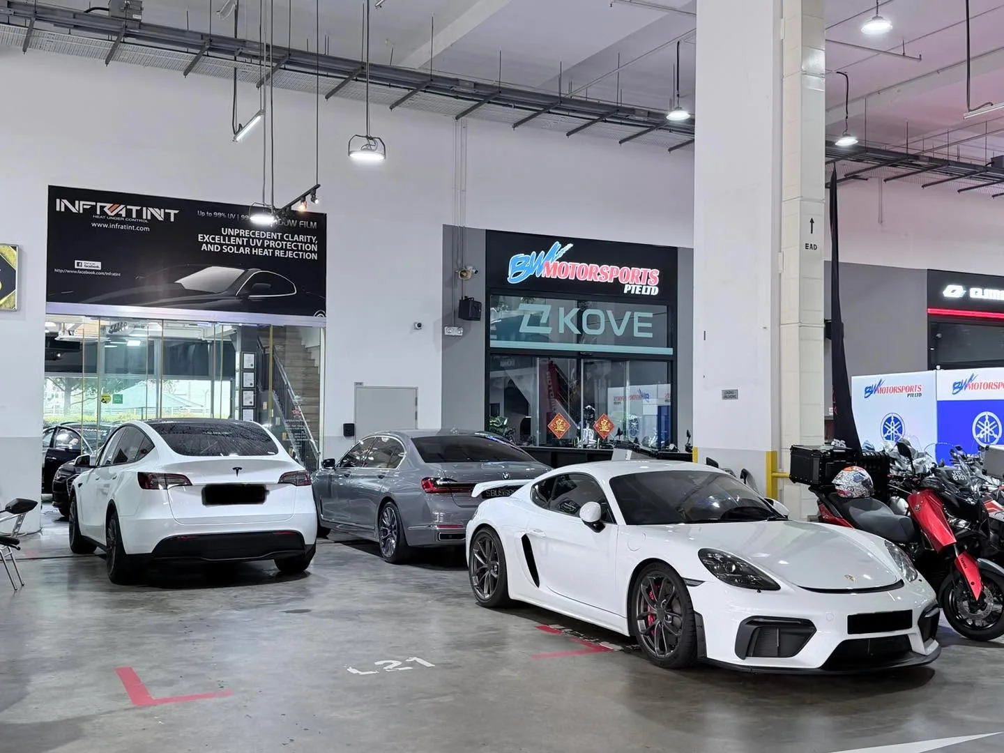 A white Porsche 911 sports car in the foreground, a white Tesla Model Y saloon behind it, and other vehicles at the Infratint workshop.