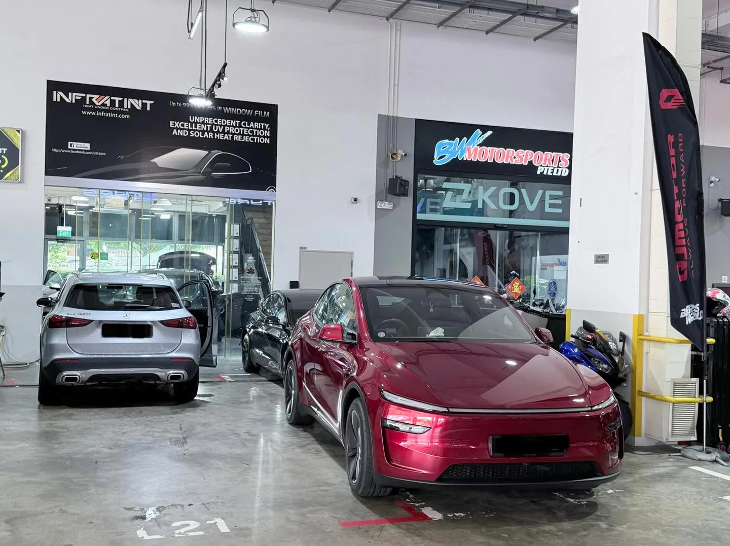 A white BMW hatchback and red Tesla sedan in the workshop with signage visible.