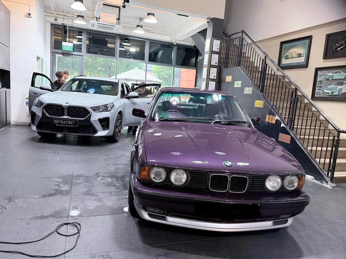 A white BMW hatchback and purple BMW saloon at an indoor facility with people nearby.