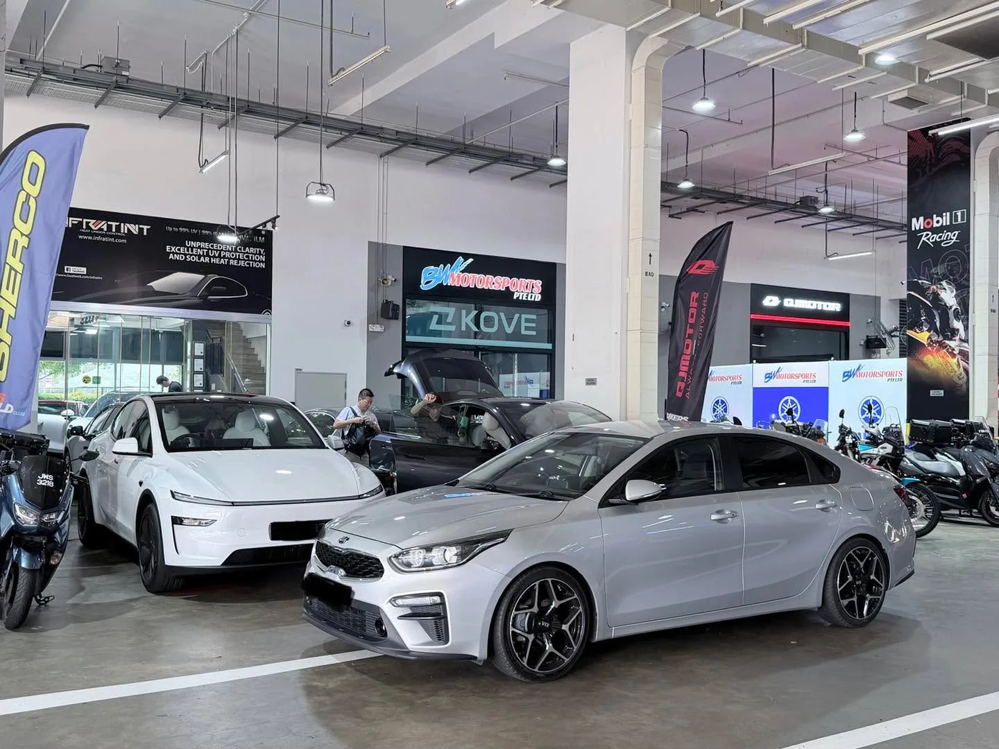 A white Tesla sedan and a dark grey Kia hatchback in the workshop, with a technician working on the Kia.