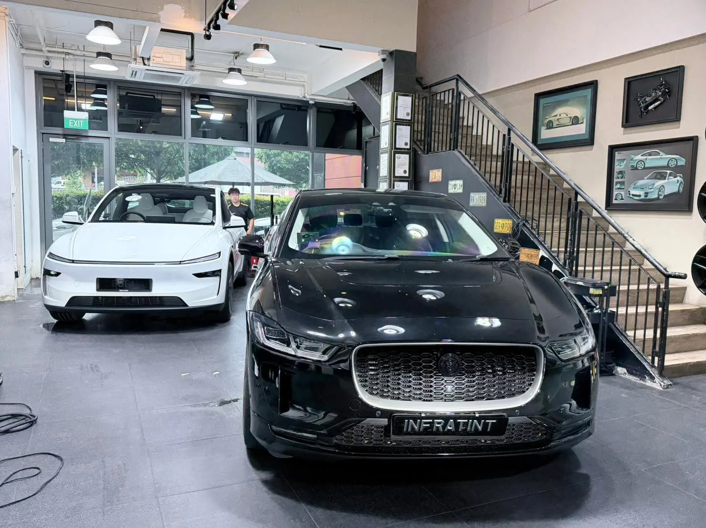 A black Jaguar electric crossover facing the camera in the showroom with a white Tesla sedan behind it.