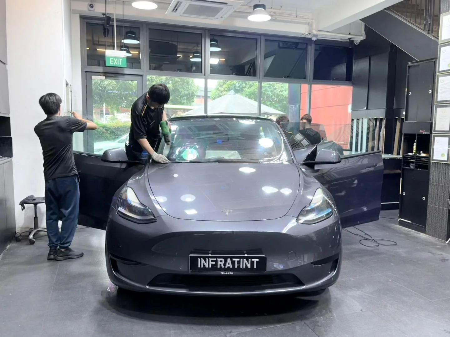 Dark grey Tesla being tinted by multiple technicians inside the workshop.
