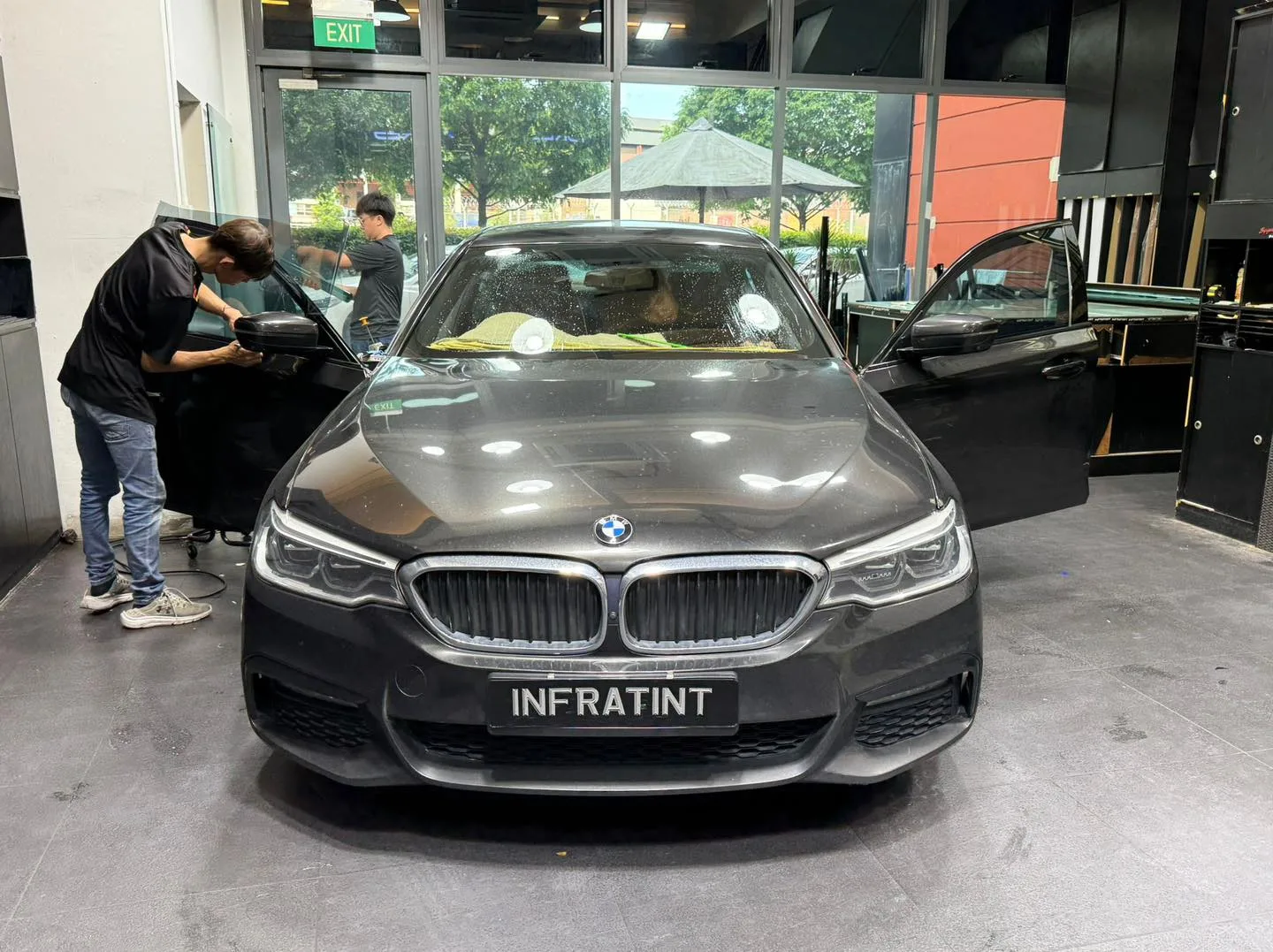 Dark grey BMW sedan with doors open, technicians tinting windows.