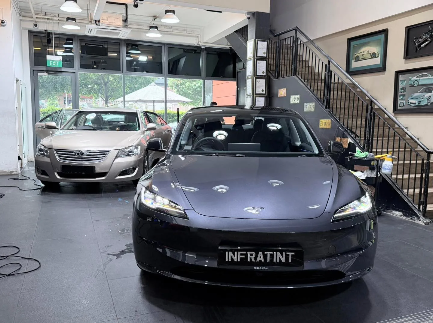 A black Tesla sedan in the foreground with a beige Toyota sedan behind it in the workshop.