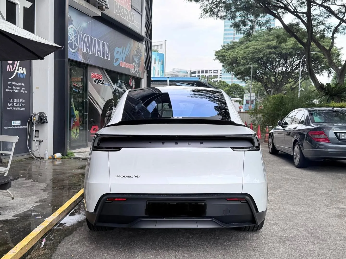 A white Tesla Model Y is parked outside a workshop, viewed from the rear.