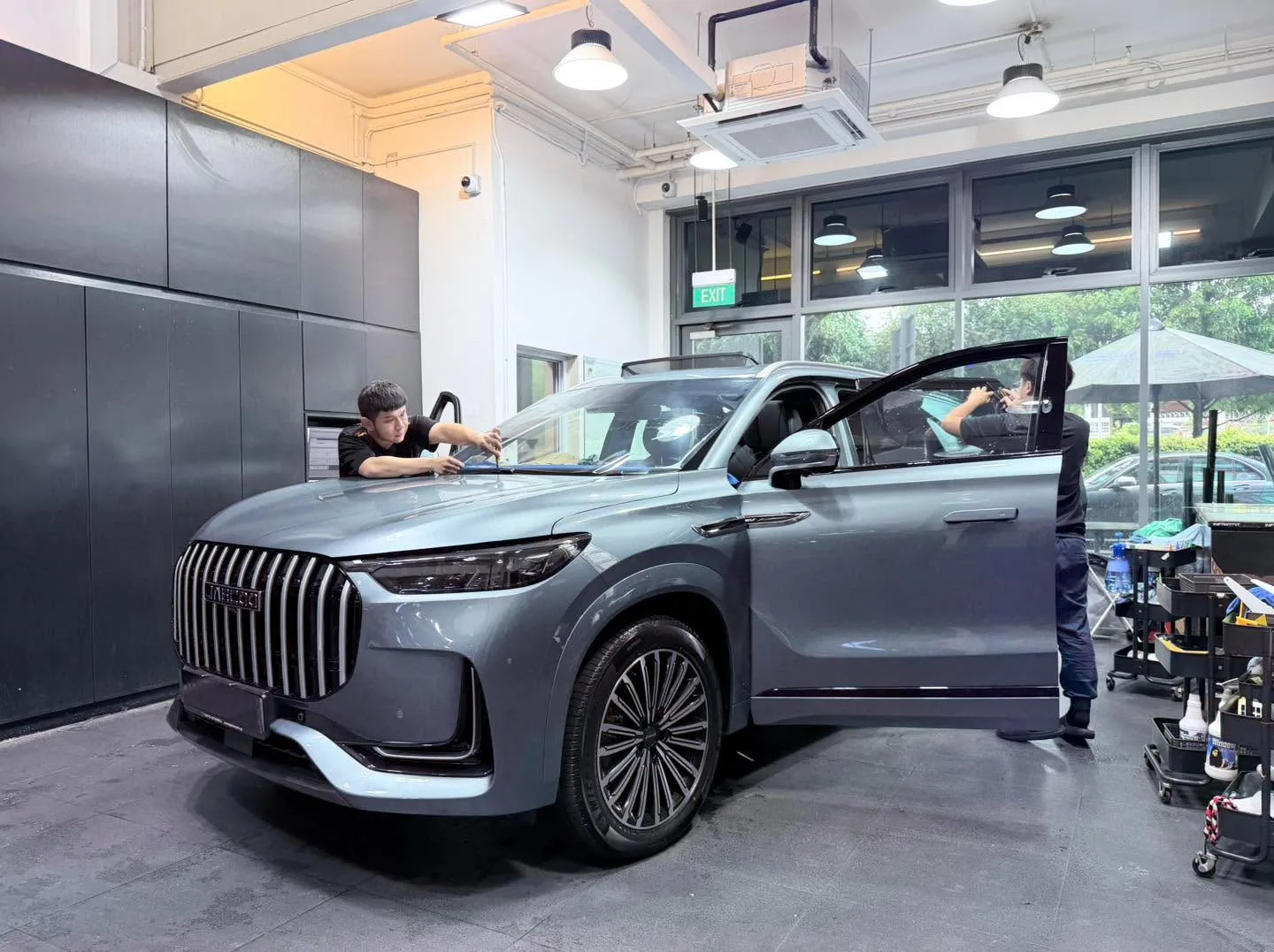 Two technicians applying window film to the roof and side windows of a grey SUV inside the workshop.