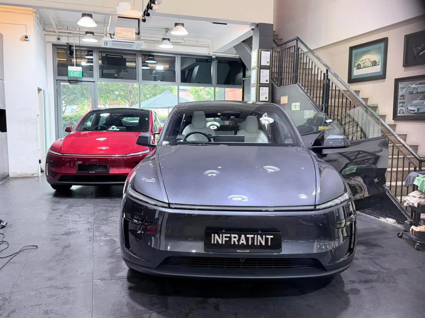 A white Tesla Model Y and a red Tesla Model Y in the workshop with glass doors visible.