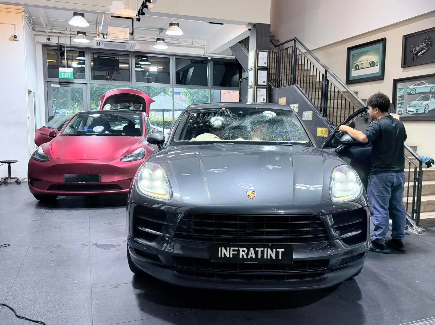 A grey Porsche Macan and a red Tesla Model Y indoors, with a technician working.