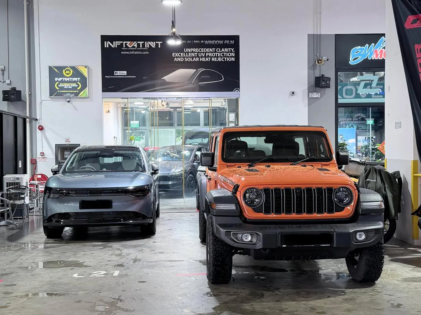 An orange Jeep Wrangler with distinctive seven-slot grille and a dark sedan parked in the workshop.