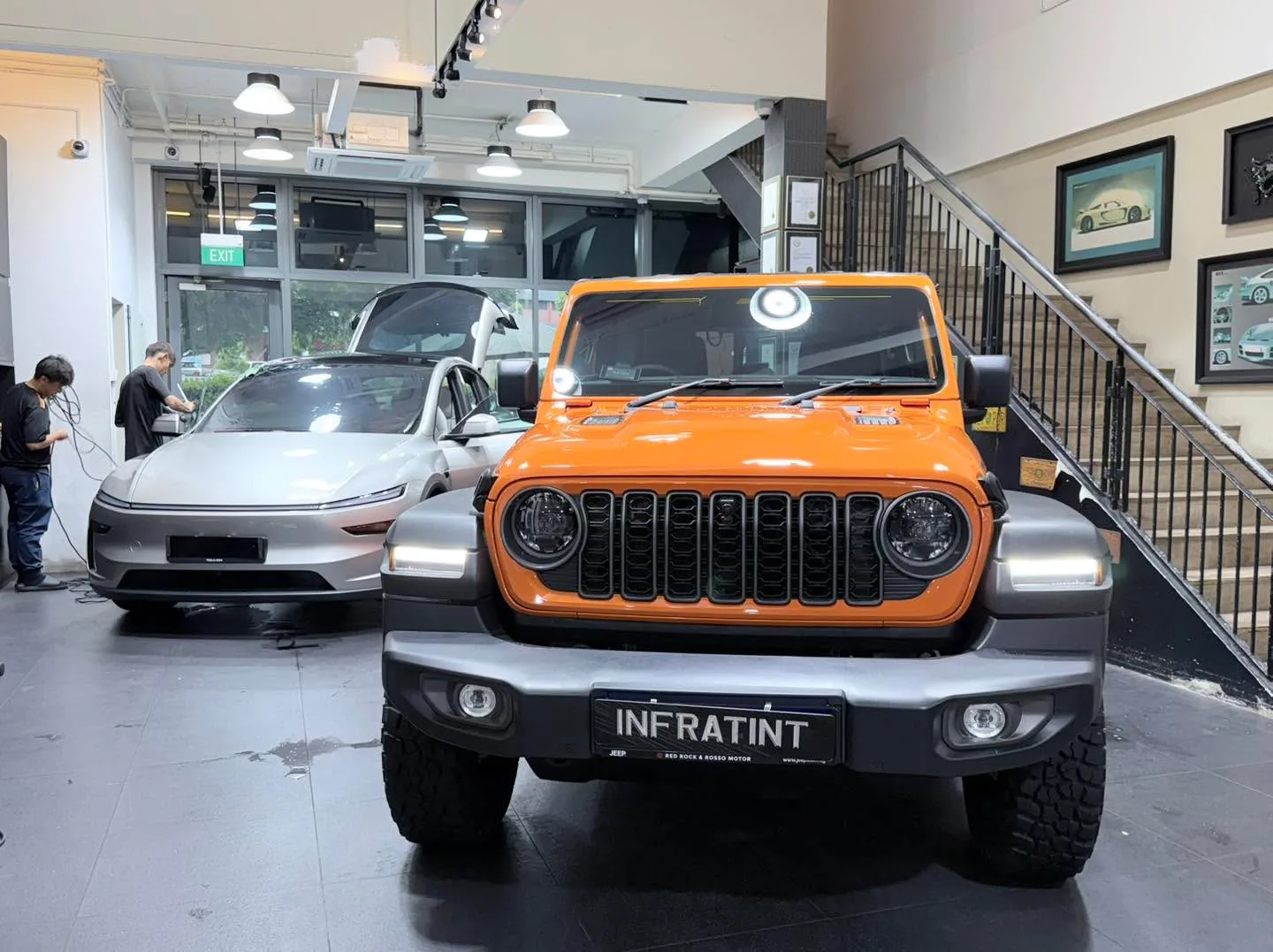 An orange Jeep Wrangler and a silver Tesla sedan parked in the workshop with staff present.
