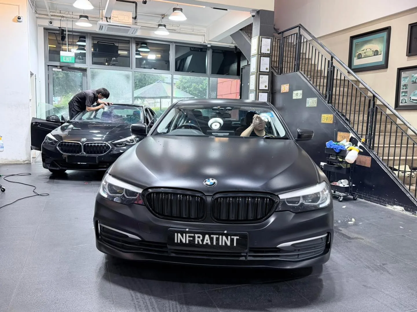 A black BMW sedan and a dark Mercedes-Benz SUV indoors with technicians working.