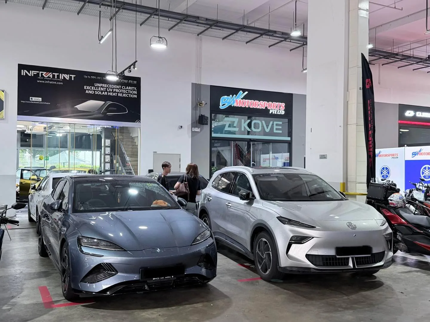 A grey saloon and a white electric SUV parked indoors in a workshop with tinted windows.
