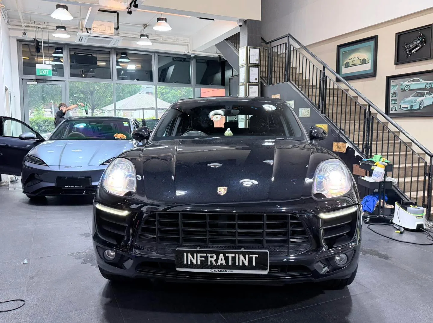 A black Porsche SUV parked front-on in an indoor workshop with stairs and artwork visible.