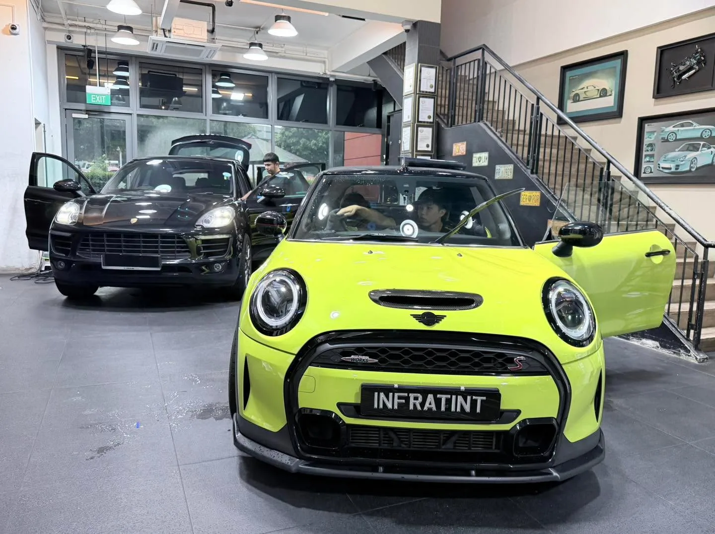A bright yellow Mini Cooper electric hatchback parked front-on indoors with a black Porsche behind.