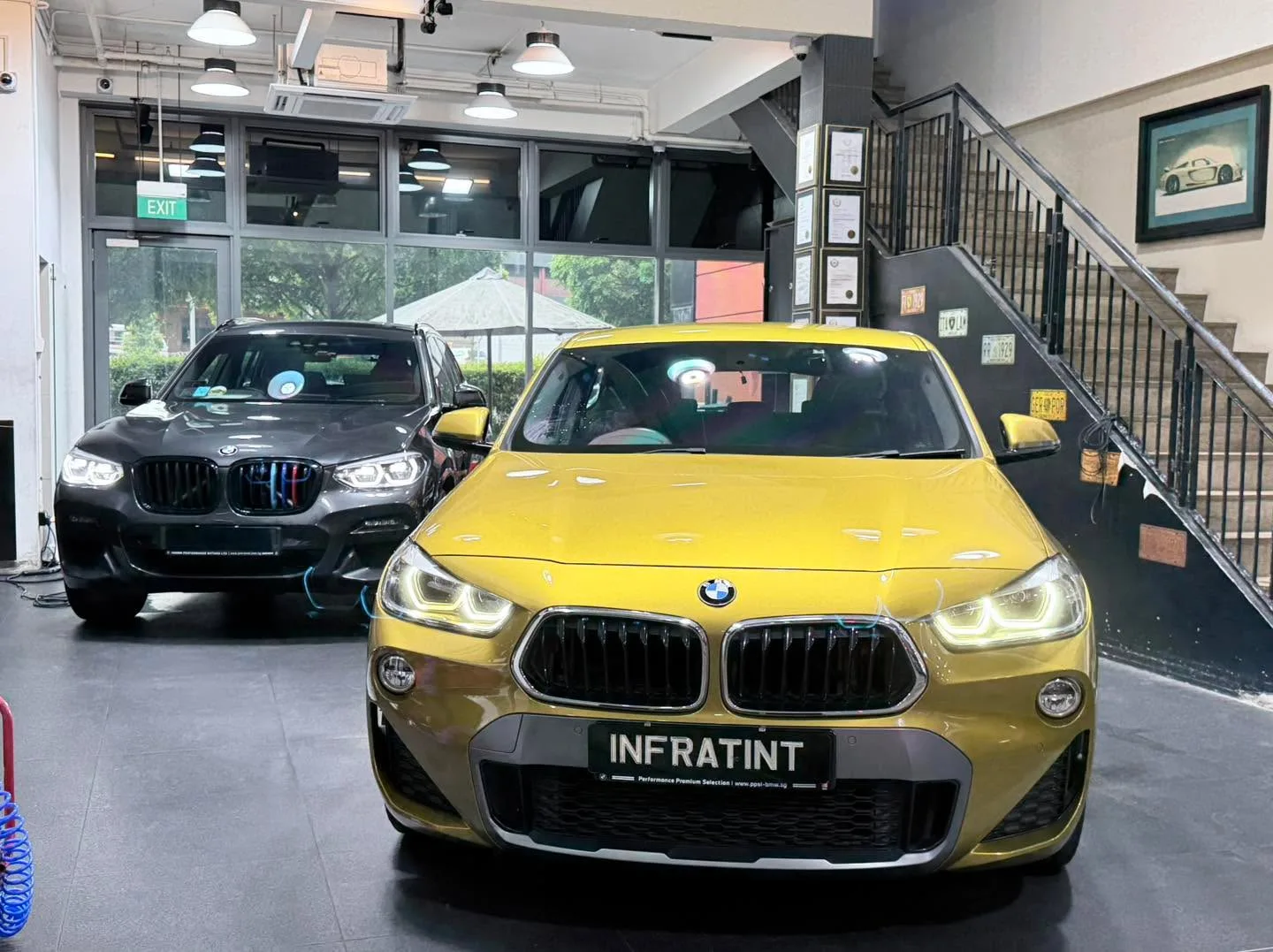 A gold BMW saloon and a black BMW X-series parked indoors by large glass windows.