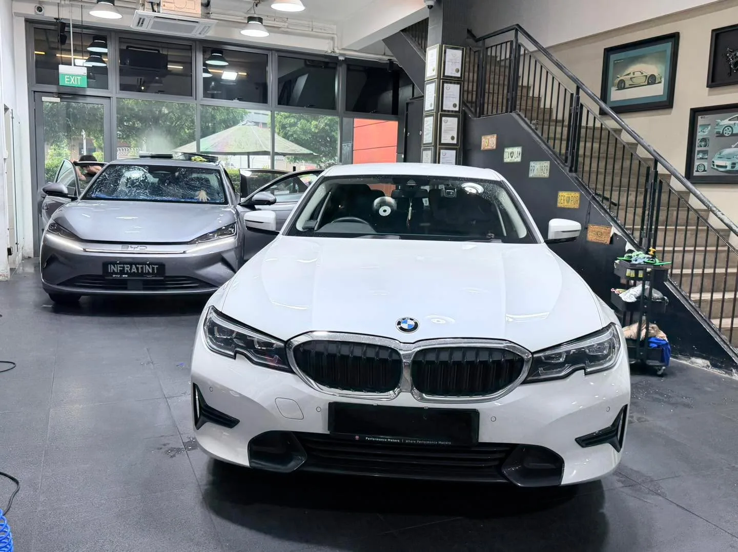 A white BMW saloon parked front-on in the workshop with open door, stairs and signage visible.