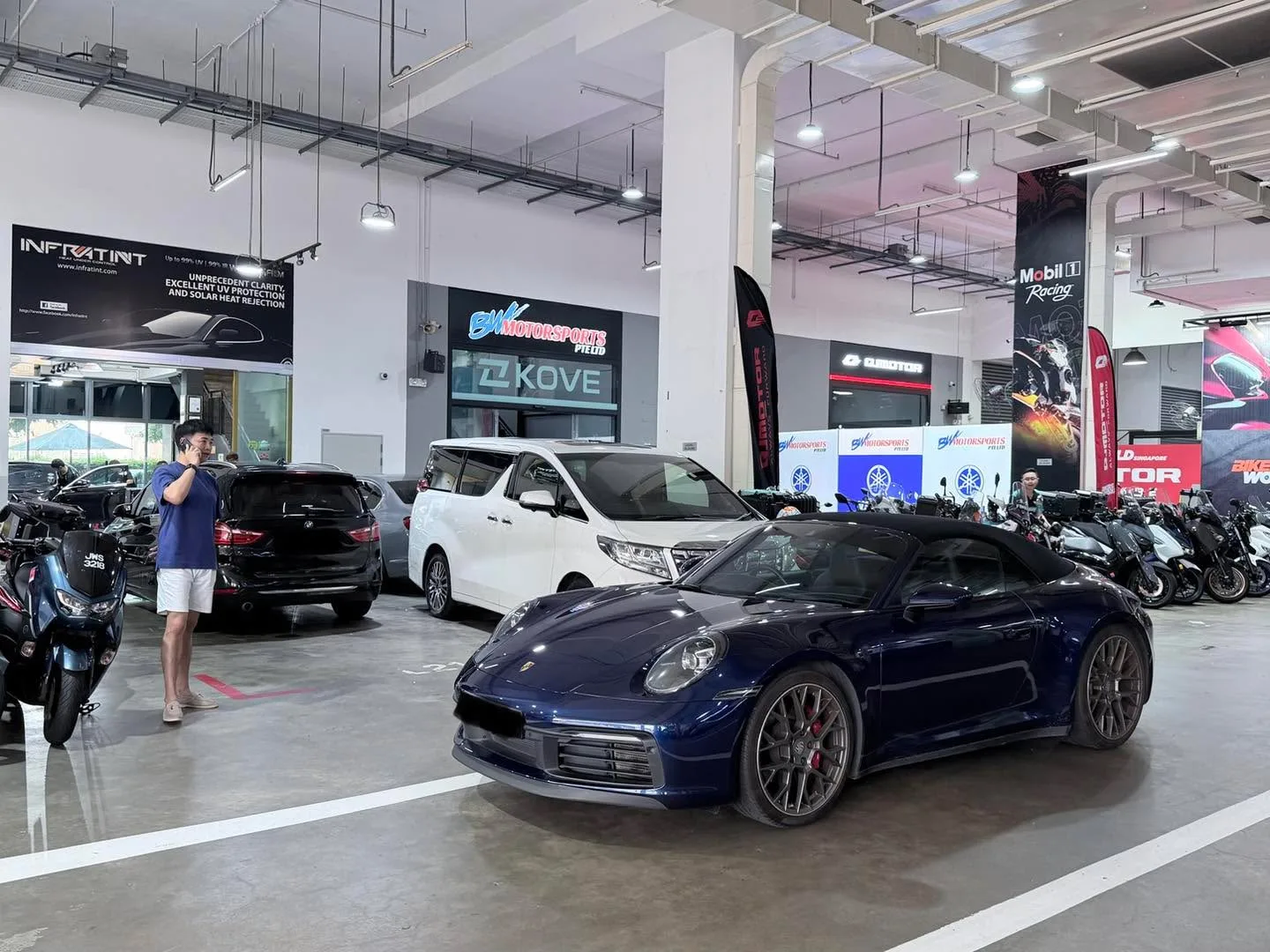 A blue Porsche 911 convertible in a workshop with motorcycles and other cars visible.