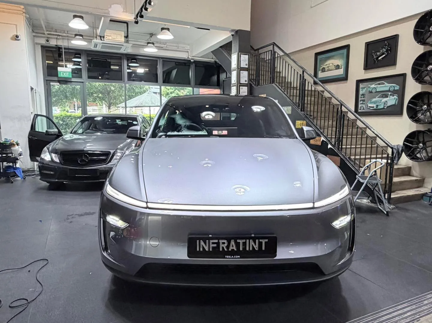 A silver Tesla sedan viewed from the front inside a showroom with black stairs and artwork on the walls.