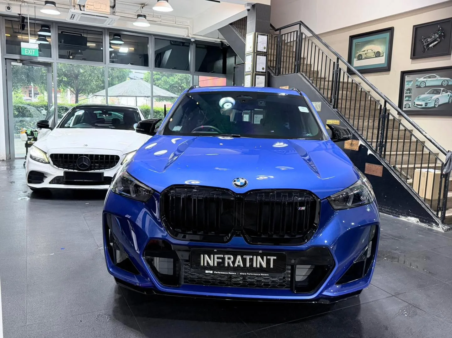 A bright blue BMW X1 M135i SUV viewed from the front inside the workshop with a white Mercedes-Benz behind it.