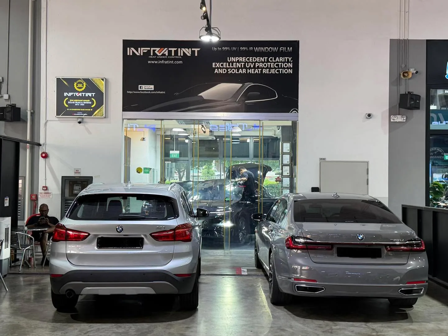 A white BMW X1 and a silver BMW sedan parked side by side in the workshop, viewed from behind.