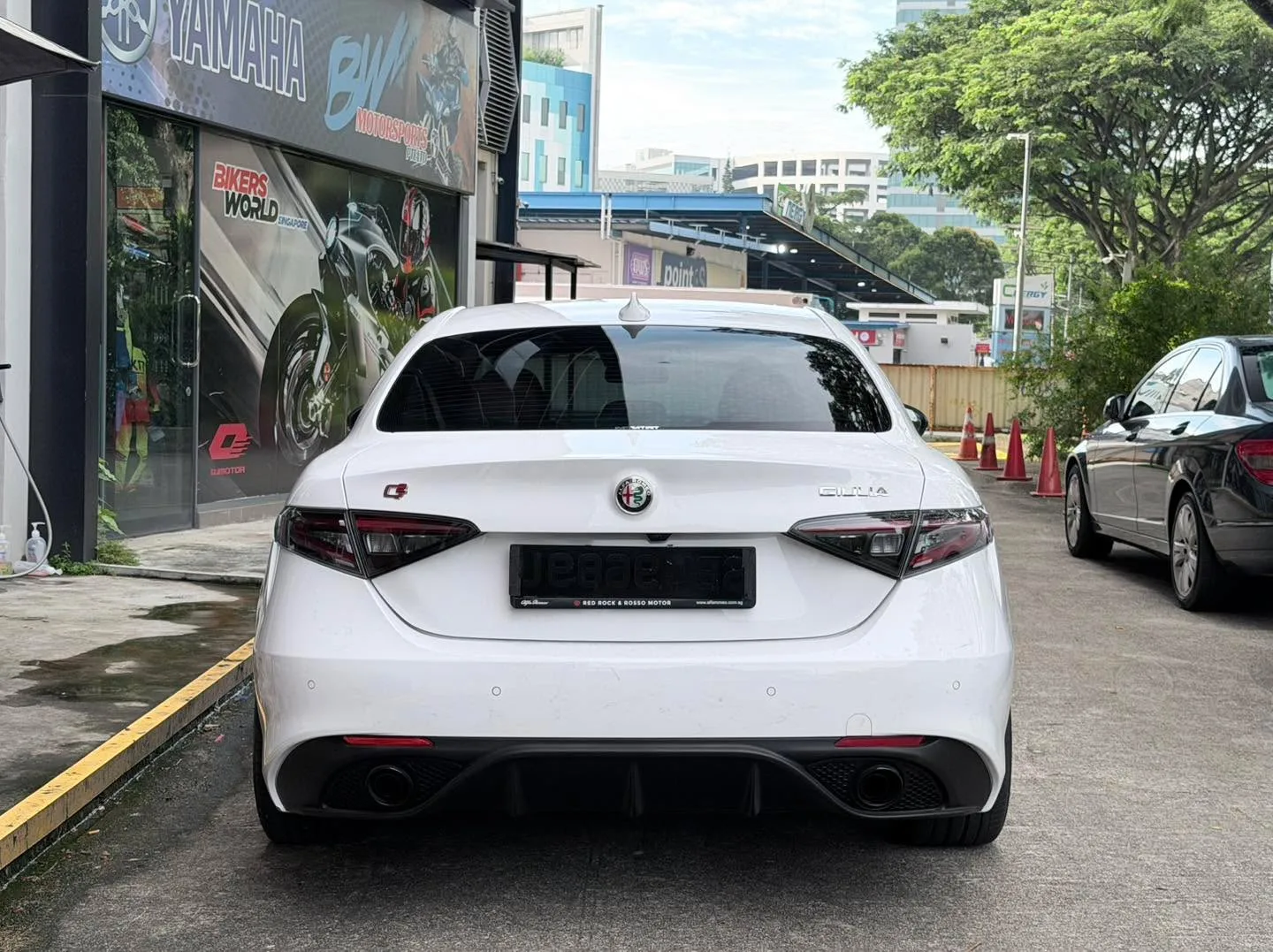 White Alfa Romeo Giulia from behind, parked on a street with motorcycle shop visible nearby.