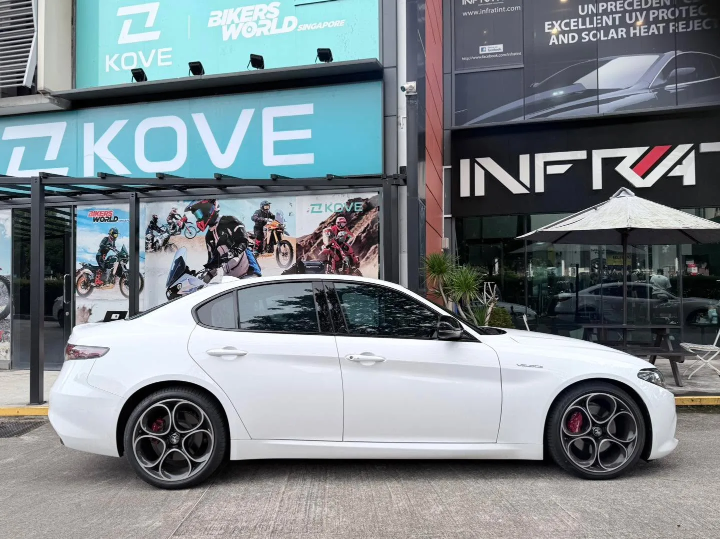 White Alfa Romeo Giulia in profile, parked outside storefronts with KOVE and Infratint signage.