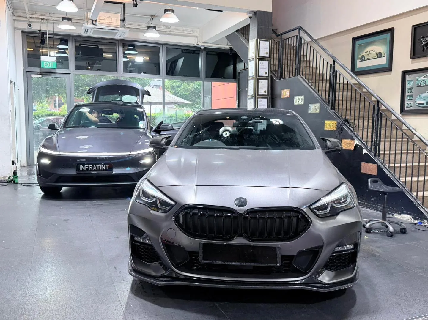 A black BMW sedan and a silver hatchback parked in a workshop with equipment nearby.