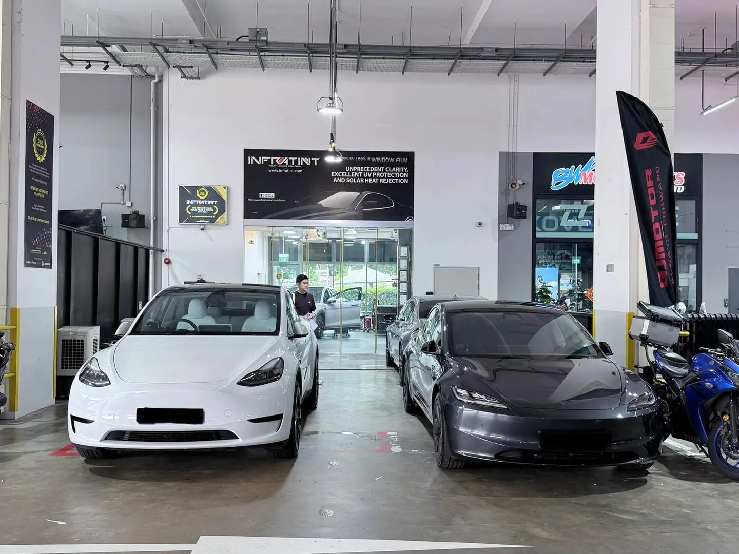 A white Tesla sedan and a dark grey Tesla sedan parked side by side in the workshop.