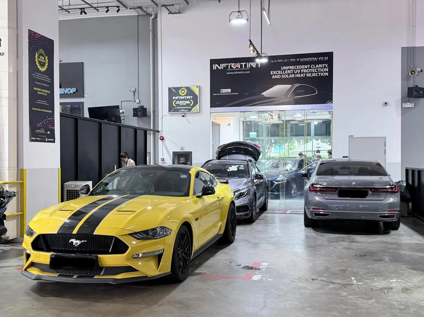 A yellow Ford Mustang with black stripes parked between a dark blue sedan and a silver sedan in the workshop.