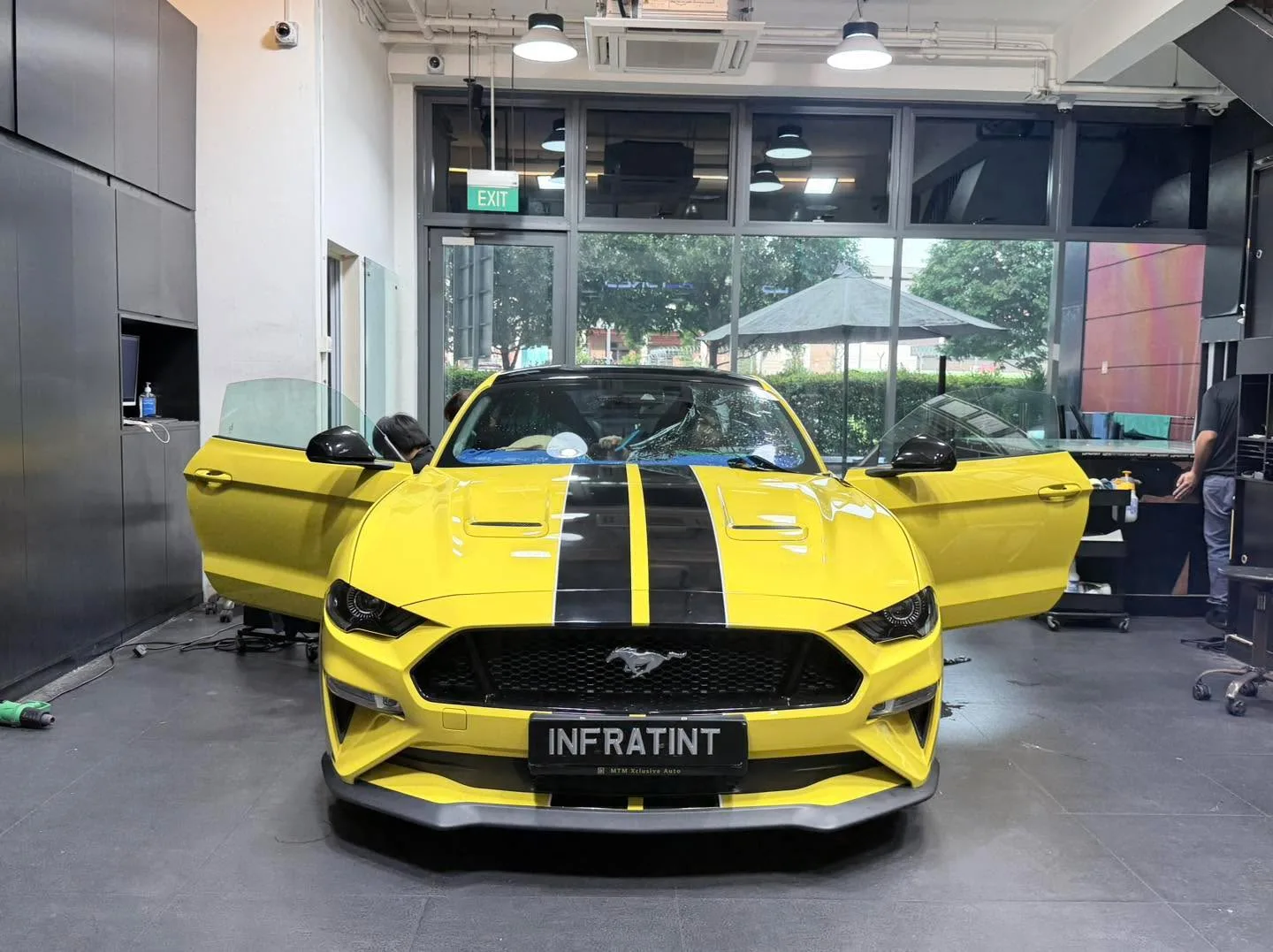 A yellow Ford Mustang 5.0 with black stripes, viewed from the front with doors open, in the workshop.