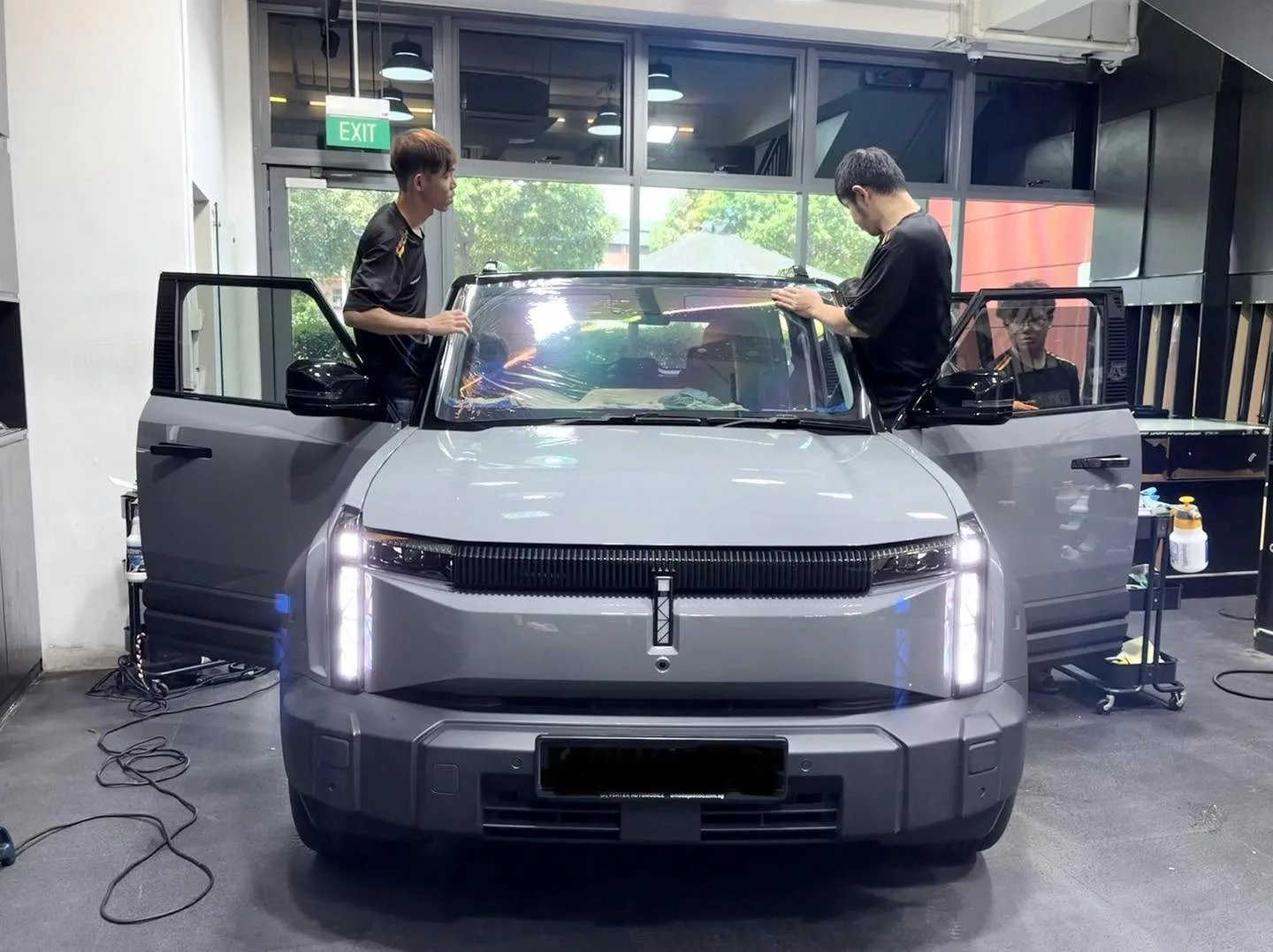 A grey electric SUV with its doors open, two technicians applying film to the front windscreen.