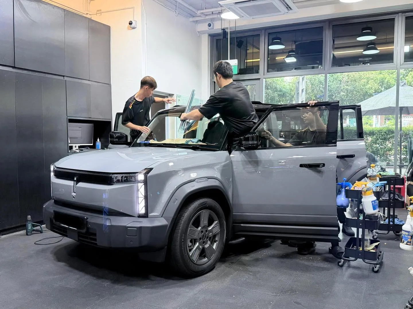 A grey electric SUV with its rear hatch open, technicians working on the rear windows.