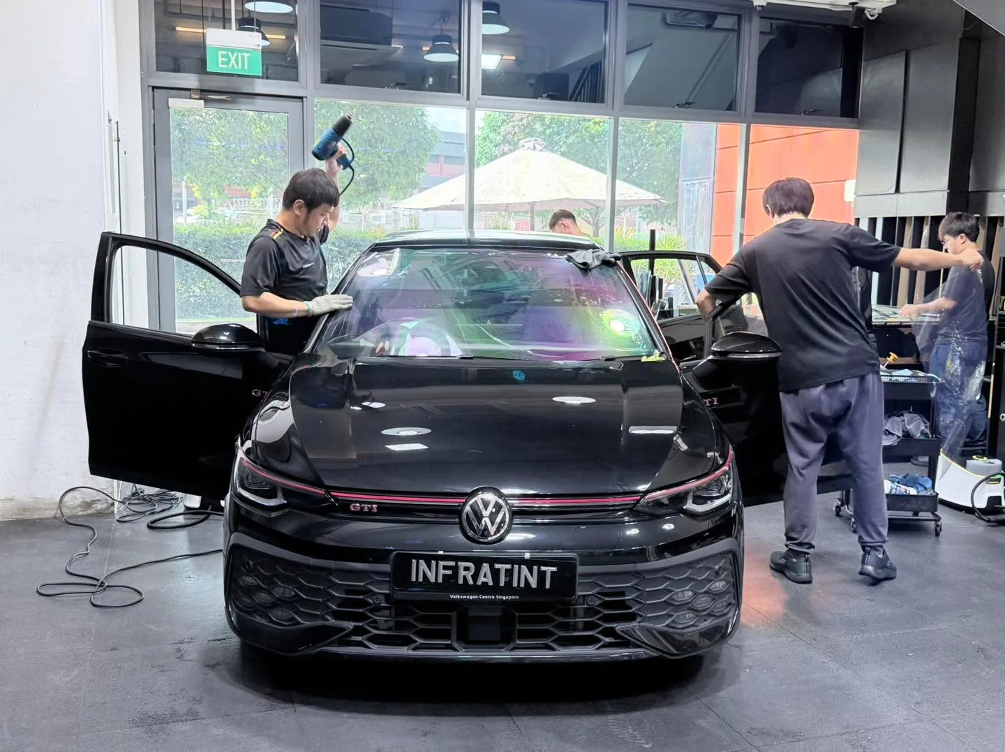 A black Volkswagen GTI with doors open, technicians applying tint film to the windscreen and side windows.