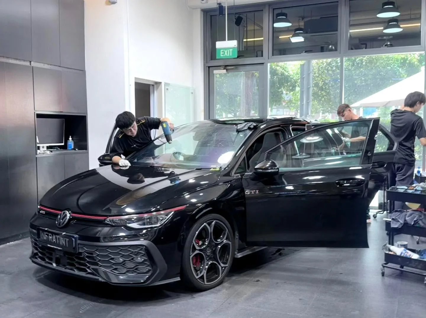 A black Volkswagen GTI with doors open, technicians applying tint to the side windows.