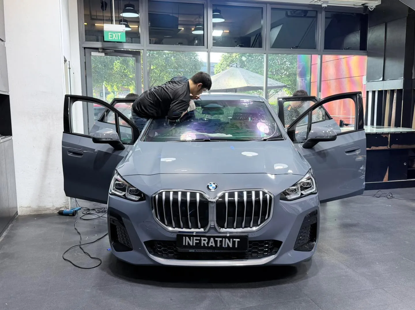 A grey BMW with its doors open, a technician applying tint to the windscreen.