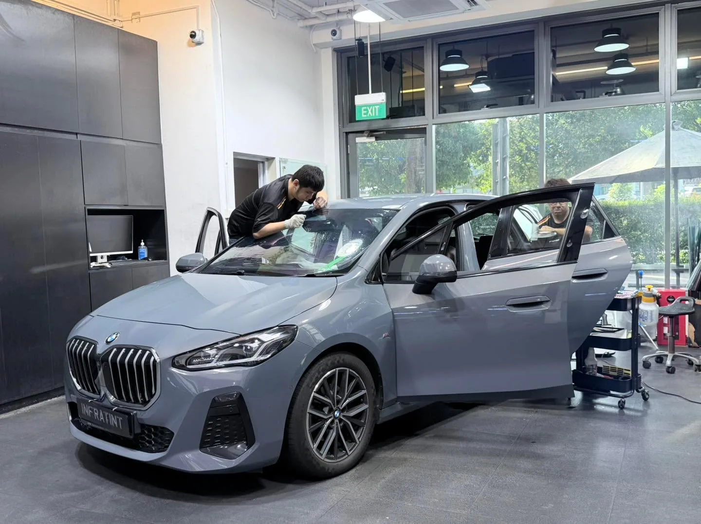 A grey BMW with doors open, a technician working on the windscreen tint.